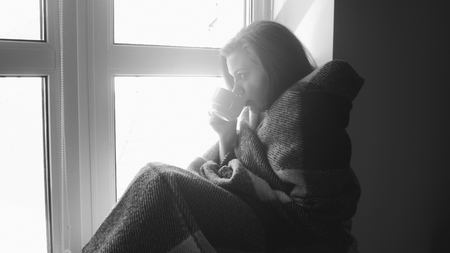 Black and white photo of young woman in plaid drinking tea at window