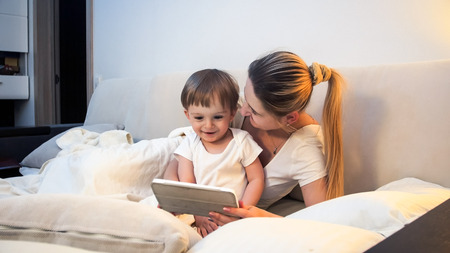 Happy smiling mother and toddler lying in bed and watching movie on digital tablet