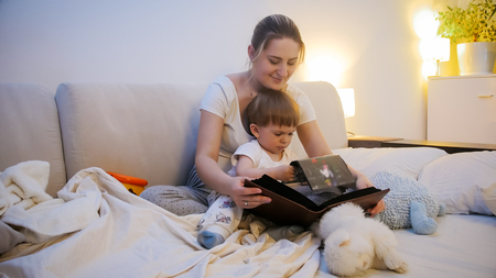 Cute toddler boy watching family photo album with his mother in bed at nightの写真素材
