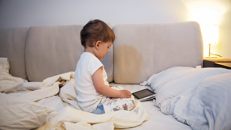 Adorable toddler boy in pajamas sitting o nbed at night and watching cartoons on digital tabletの写真素材