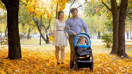 Happy smiling family walking with toddler son sitting in pram at autumn parkの写真素材