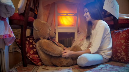 Beautiful smiling girl sitting in blanket tent and playing with teddy bearの写真素材