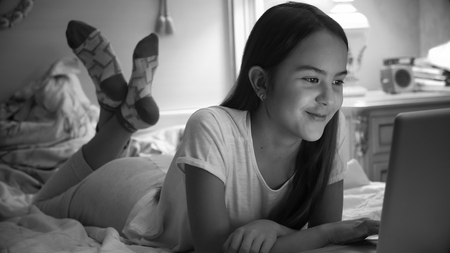 Black and white portrait of cute smiling girl in pajamas lying on bed with laptopの写真素材