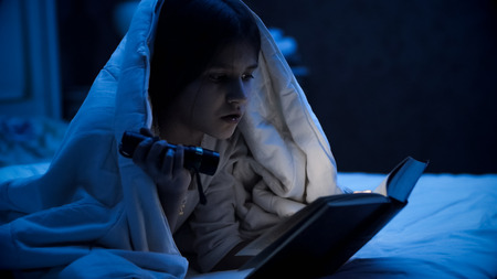 Closeup portrait of little girl lying in bed and reading book with flashlightの写真素材