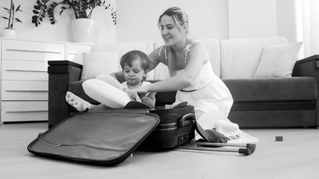 Cute smiling toddler boy sitting in suitcase while mother packing things for travelingの写真素材