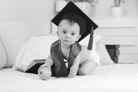 Black and white image of genius boy in graduation hat sitting on bed with apple and looking in cameraの写真素材