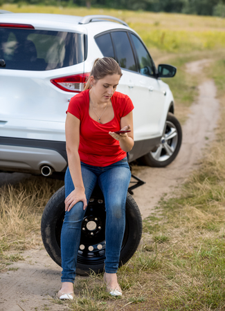 Young woman with broken car sittign on spare wheel and calling auto service for helpの写真素材