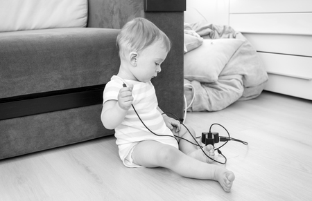 Black and white image of little baby boy sitting alone at room and pulling electric cables and wiresの写真素材