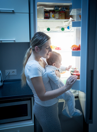 Portrait of young mother looking for food in refrigerator at night to feed her little baby boyの写真素材