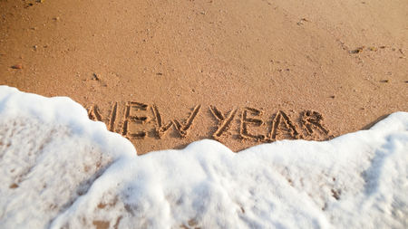 Closeup image of sea waves rolling over words New Year written on wet sand on the beach. Concept of...