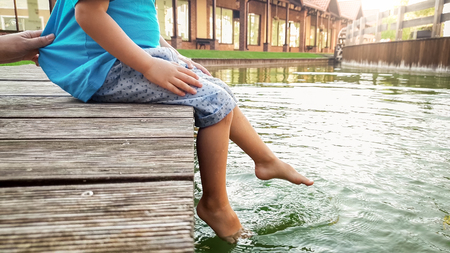 Closeup image of barefoot little toddler boy sitting on wooden bridge at lake and splashing water with his feetの写真素材