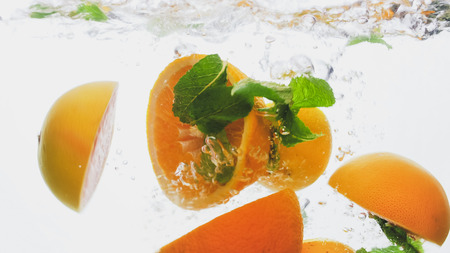 Closeup photo of cut fresh juicy oranges with mint leaves floating in clear water against white backgroundの写真素材