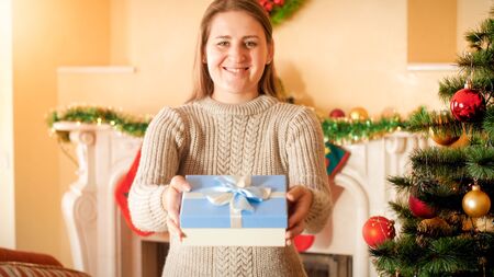 Portrait of happy smiling young woman in wool sweater holding Christmas gift in box and showing it...