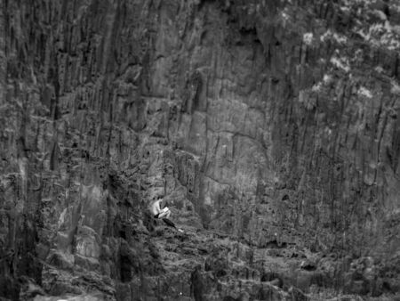 Black and white image of young lonely man sitting on the cliff at sea shore and reading bookの写真素材