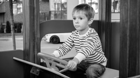 Black and white portrait of cute toddler boy palying in big wooden car on children playground at parkの写真素材