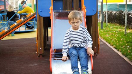 Sad and upset little lonely boy sitting on slide at playgroundの写真素材
