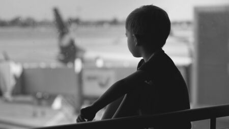 Black and white upset little boy sitting in airport terminal and looking on big airplanesの写真素材