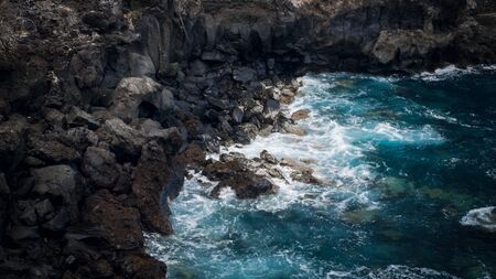 Seascape of blue ocean waves rolling over black volcanic rocks and cliffs on the shoreの写真素材