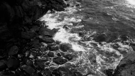Black and white photo of ocean waves rolling and breaking on sharp cliffs and rocks on the beach