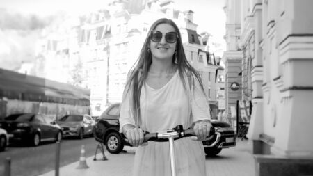 Black and white portrait of beautiful smiling woman in sunglasses riding on electric scooter on city...
