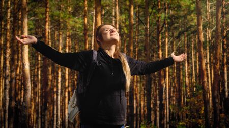 Portrait of beautiful smiling female tourist with backpack with outstretched hand looking on bright sun in pine tree forest. Concept of freedom and harmony with nature.の写真素材