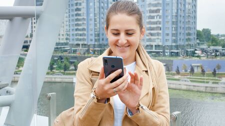 Portrait of beautiful smiling young woman walking on the modern glass bridge and using smartphone