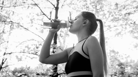 Black and white portrait of beautiful young woman in sports outfit drinking water from bottle after exercising in parkの写真素材