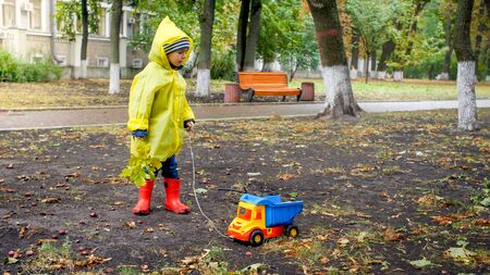 Little toddler boy in raincoat playing with toy truck at park on cold rainy dayの写真素材