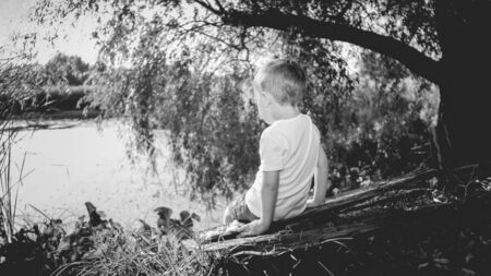Black and white image o flittle boy sitting on the wooden log at river and looking on the sunsetの写真素材