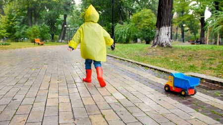 Cute little boy in yellow raincoat running with toy truck under rain at parkの写真素材