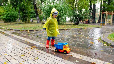 Cute little boy in raincoat and rubber boots blaying with toy truck in big puddle at parkの写真素材
