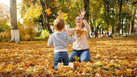 Happy laughing mother with little son throwing autumn leaves at parkの写真素材