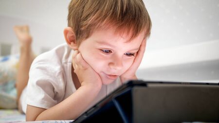 Portrait of smiling little boy in pajamas lying in bed and using digital tablet computerの写真素材