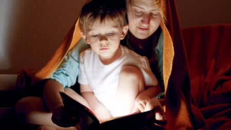 Portrait of cute little boy with young mother covering in blanket and reading a book with flashlightの写真素材