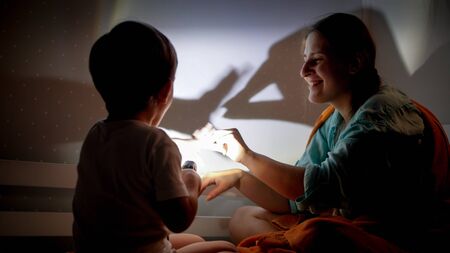 Portrait of smiling young mother and little boy playing with flashlight and shadows on wall at night