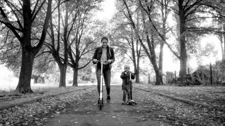 Black and white photo of young mother with little son riding kick scooter on long alley at autumn parkの写真素材