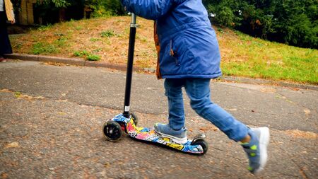 Closeup photo of little boy standing and riding on kick scooter at autumn park