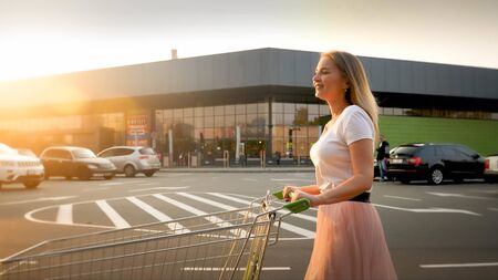 Beautiful smiling blonde girl pushing shopping cart while walking on car parking at shopping mall