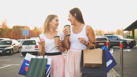 Toned photo of two women with paper bags talking and smiling while walking on car parking at shopping mallの写真素材