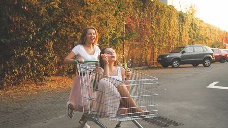 Toned photo of two happy girls riding in shopping cart and blowing soap bubbles on car parkingの写真素材