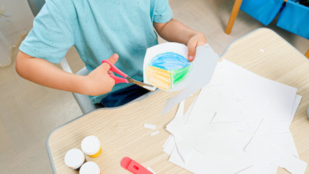 Closeup of little smart boy cutting out drawn picture with scissors. Child studying and learning behind school desk at homeの写真素材
