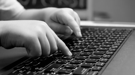 Black and white closeup photo of child pressig buttons on computer keyboard, typing message on...