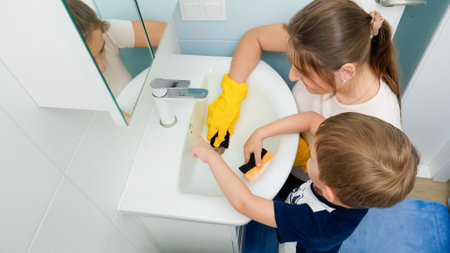 Happy smiling boy with mother wearing rubber gloves applying detergent and cleanser on sponges and washing bathroom water sink. Little boy helping parents in houseworkの写真素材