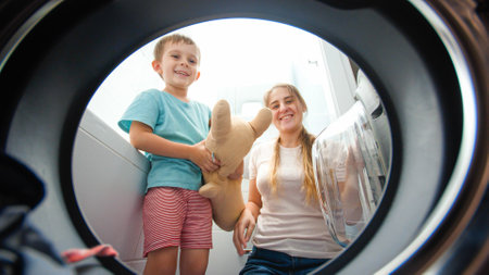 Smiling boy with young mother taking clean clothes out of washing machine in bathroom. Child helping parents washing clothes and doink choresの写真素材