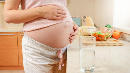 Closeup of beautiful pregnant woman taking glass of water and drinking it on kitchen. Concept of healthy lifestyle, nutrition and hydration during pregnancyの写真素材