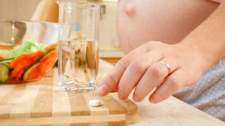 Closeup of pregnant woman taking vitamin pill and drinking glass of clear water before breakfast on kitchen. Concept of healthy lifestyle, nutrition and hydration during pregnancyの写真素材