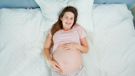 Beautiful pregnant woman in pajamas resting on bed and stroking her big belly. Concept of pregnancy,...