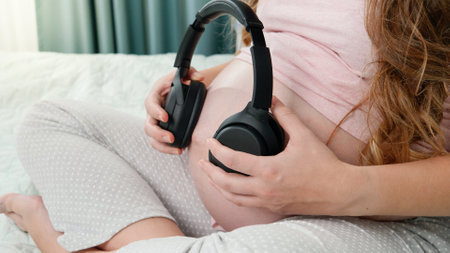 Closeup of pregnant woman sitting on bed and putting headphones on her big belly. Unborn baby...