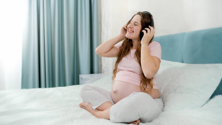 Happy smiling pregnant woman in pajamas listening to music with headphones on bed at morningの写真素材