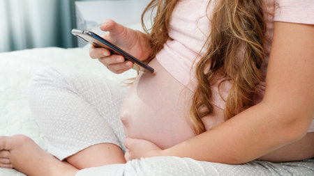 Closeup of young pregnant woman sitting on bed and browsing internet on smartphoneの写真素材